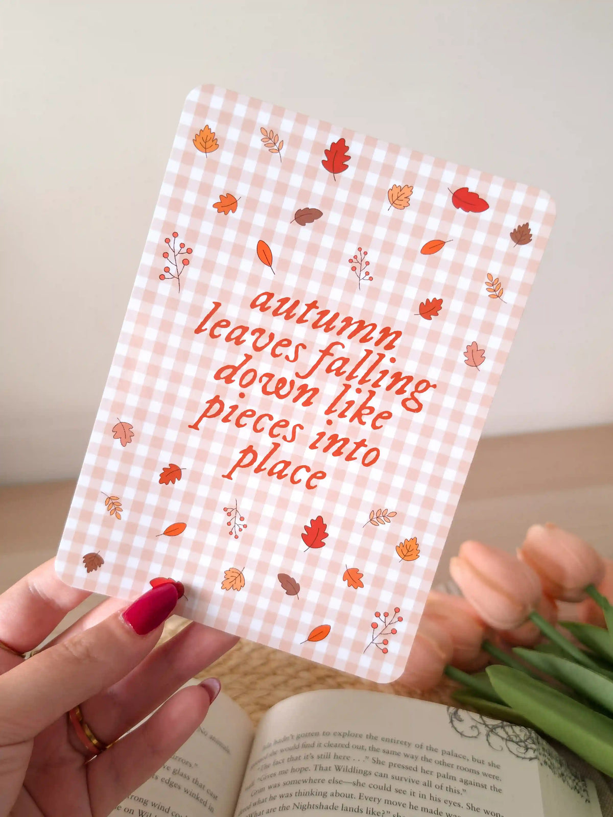 A laminated e-reader insert on a pale pink gingham background featuring the text "autumn leaves falling down like pieces into place" in orange-red cursive lettering at the centre, surrounded by illustrated autumn leaves in shades of red, orange, and brown, and small berry branches, resting on an open book on a woven rattan mat, with green eucalyptus branches visible in the upper left corner.