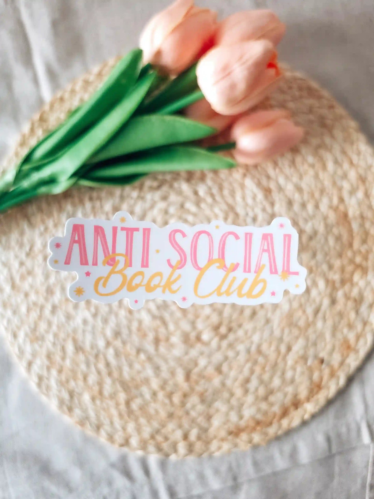 A die-cut sticker reading "Anti Social Book Club" in pink block lettering and gold cursive script, with small star details on a white background, placed on a woven rattan mat with peach roses and green leaves resting in the upper corner.