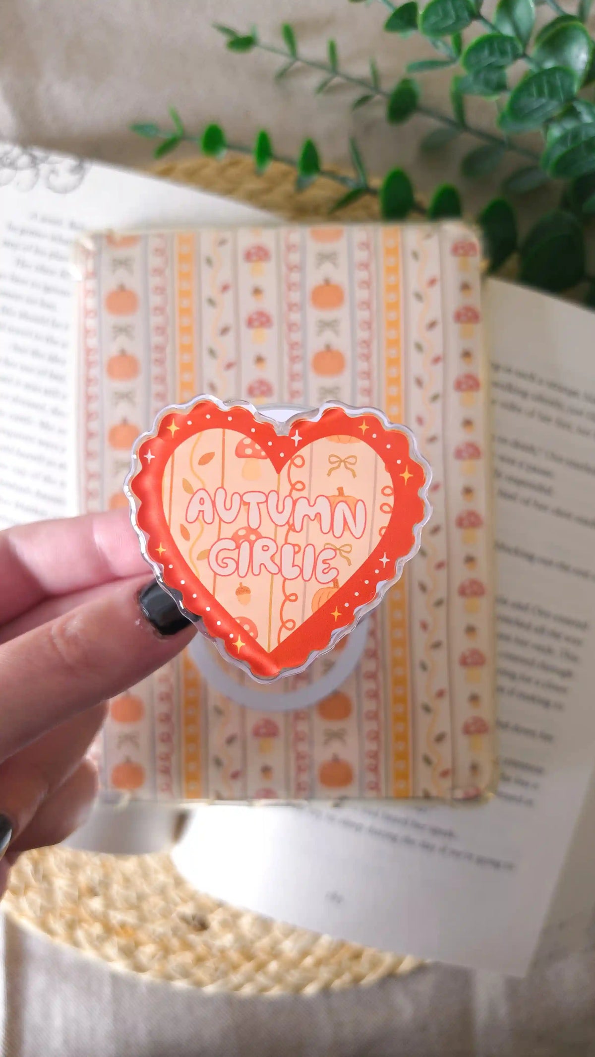 Hand holding an orange heart-shaped acrylic grip with the text “Autumn Girlie” over a pattern of pumpkins, mushrooms and bows, in front of an autumn-patterned magnetic bookmark, an open book, a green plant and a woven rattan mat.