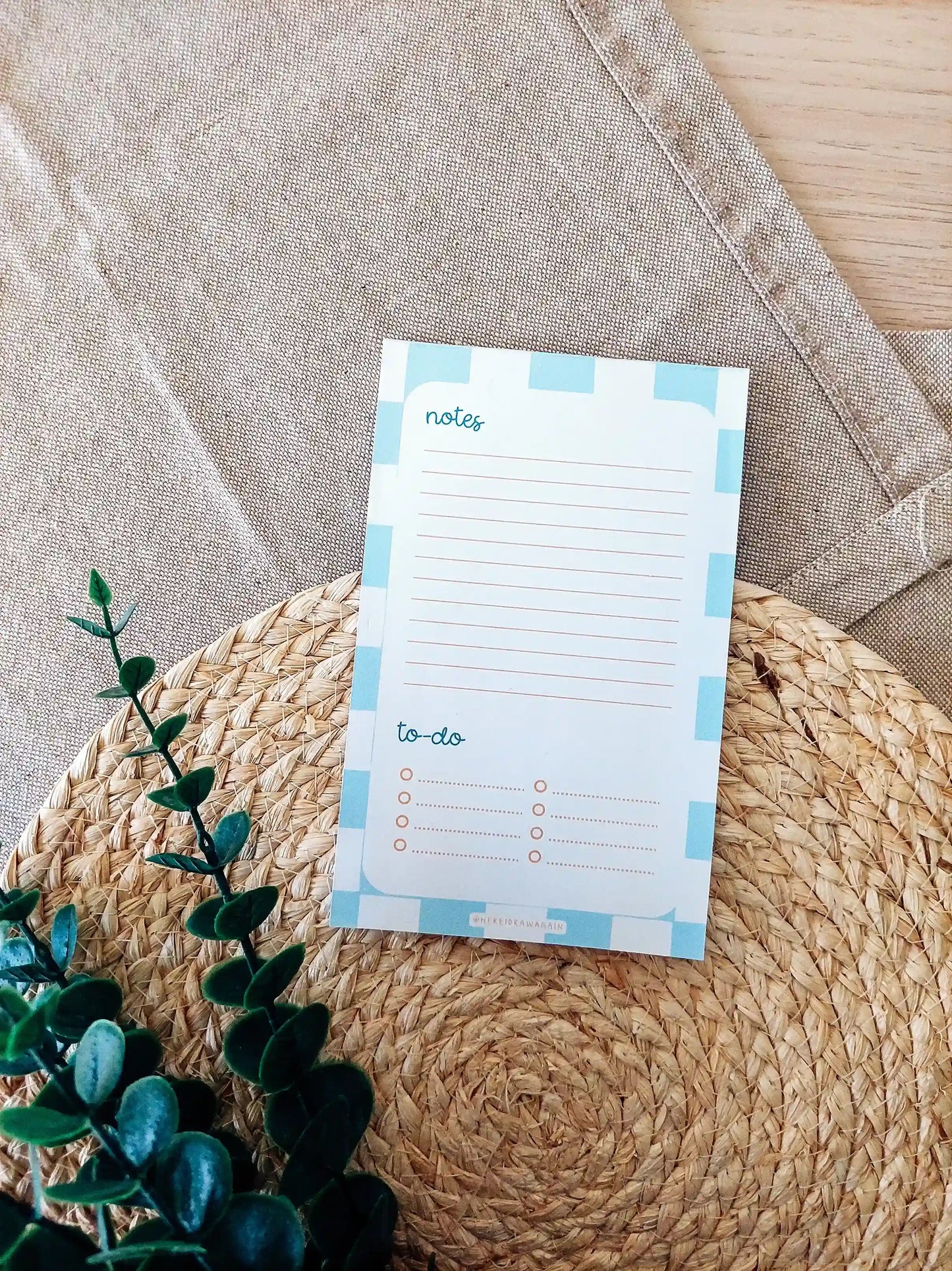 Blue checkered Notes and to-do notepad featuring a lined notes section at the top and a checklist-style to-do section at the bottom, photographed on a woven surface with decorative flowers.