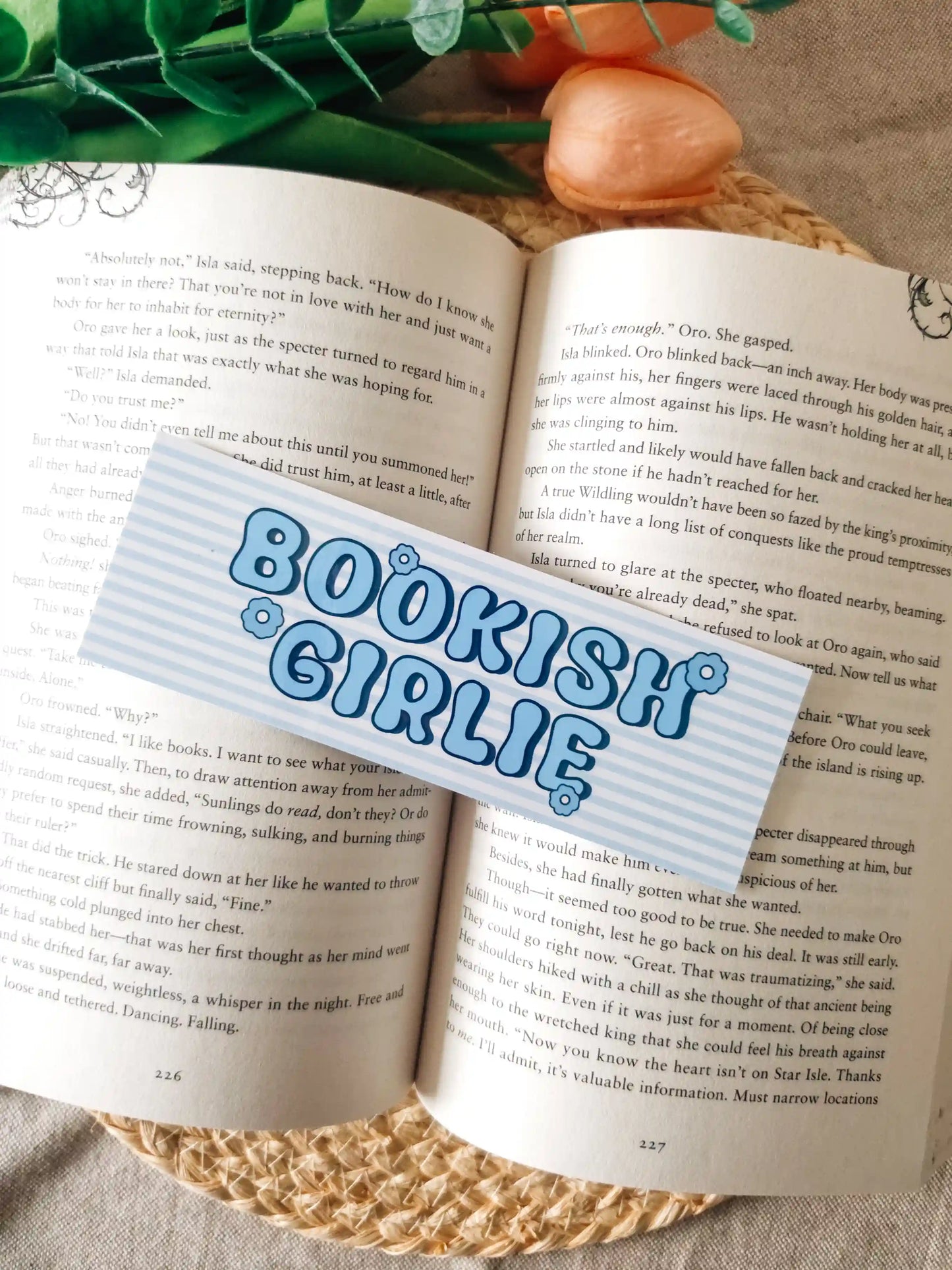 A bookmark with the words
"BOOKISH GIRLIE" in blue bubble letters, laid over an open book. The scene includes pink fake roses and green leaves at the top, set on a woven jute surface.
