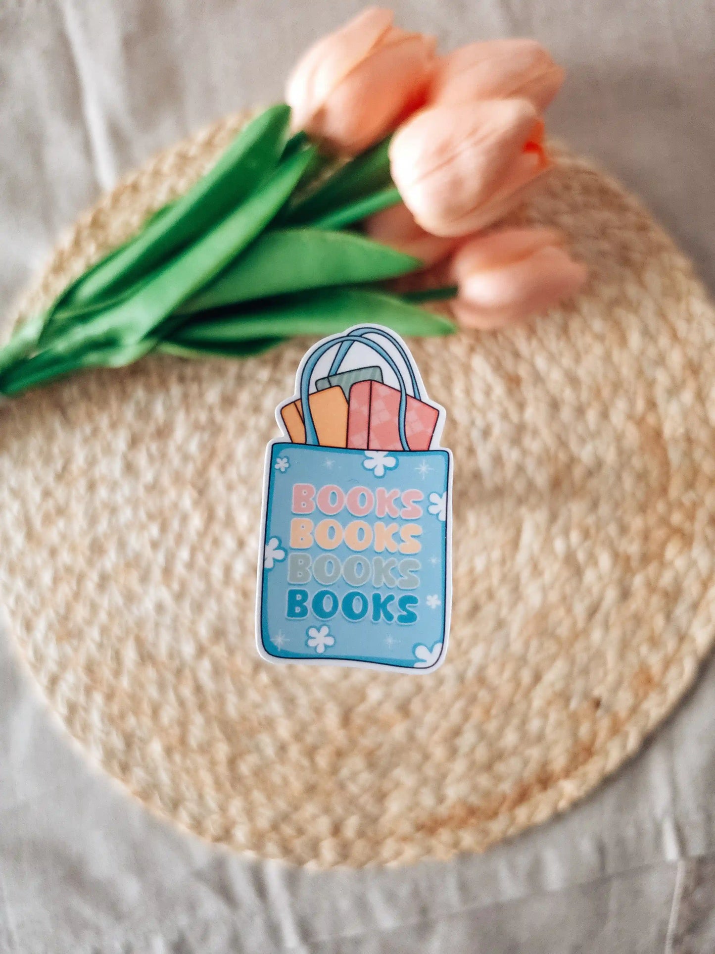 A die-cut sticker featuring a blue tote bag filled with colorful books and the text "Books Books Books Books" in pink, peach, and teal lettering, placed on a woven rattan mat. In the background, peach tulips with green leaves.