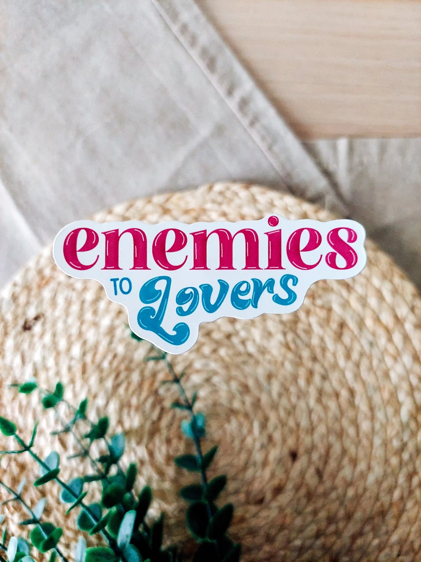 Vinyl sticker with the text "Enemies to Lovers" in pink and teal typography, photographed on neutral fabric background with woven mat and green foliage.