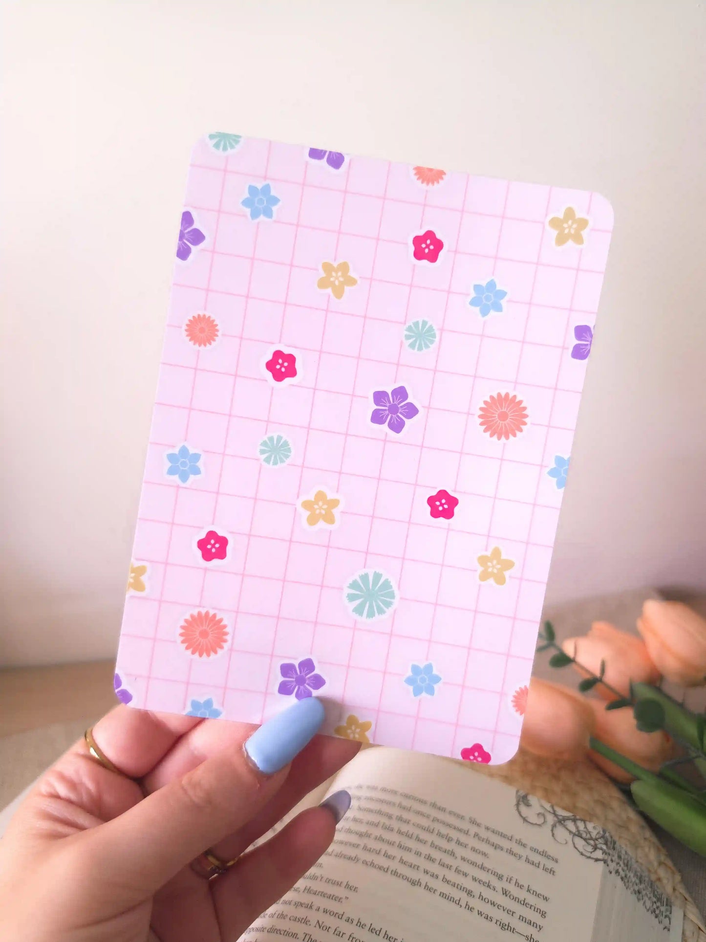 A hand holding an e-reader insert with a soft lavender grid background scattered with colorful flower illustrations in pink, blue, orange, purple, green, and yellow, placed on an open book resting on a woven rattan mat.