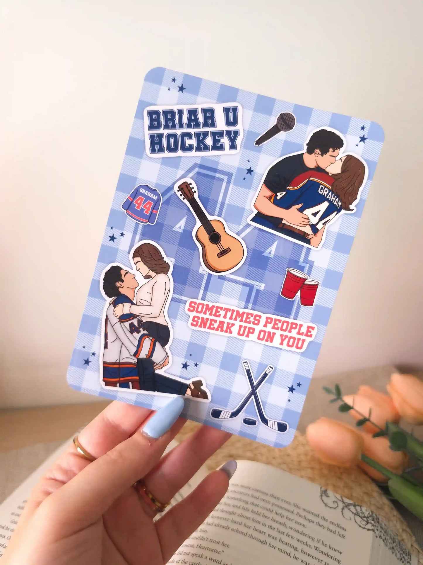 A hand holding an e-reader insert with a blue gingham background featuring illustrated elements from The Deal by Elle Kennedy, including a "Briar U Hockey" badge, a Graham #44 jersey, two illustrations of Hannah and Garrett, crossed hockey sticks, a guitar, red party cups, a microphone, and the quote "Sometimes people sneak up on you" in red lettering.