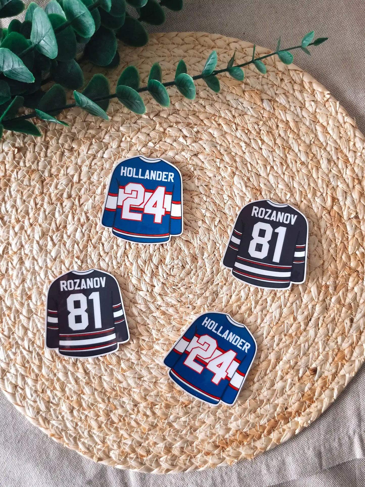 Four die-cut hockey jersey stickers arranged on a woven rattan mat, two black Rozanov #81 jerseys and two blue Hollander #24 jerseys, with green branches resting in the upper corner.