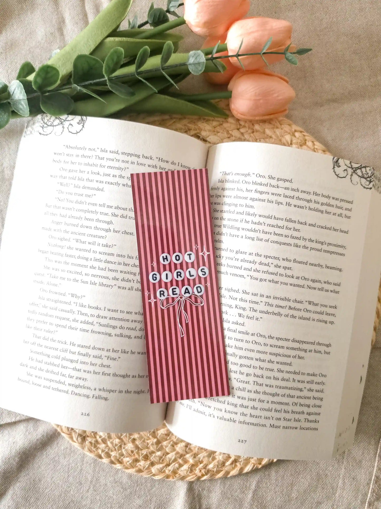 A pink striped book bookmark with the words 'HOT GIRLS READ' in white lettering style like friendship bracelets beads, placed on an open book with decorative greenery and fruit around it.