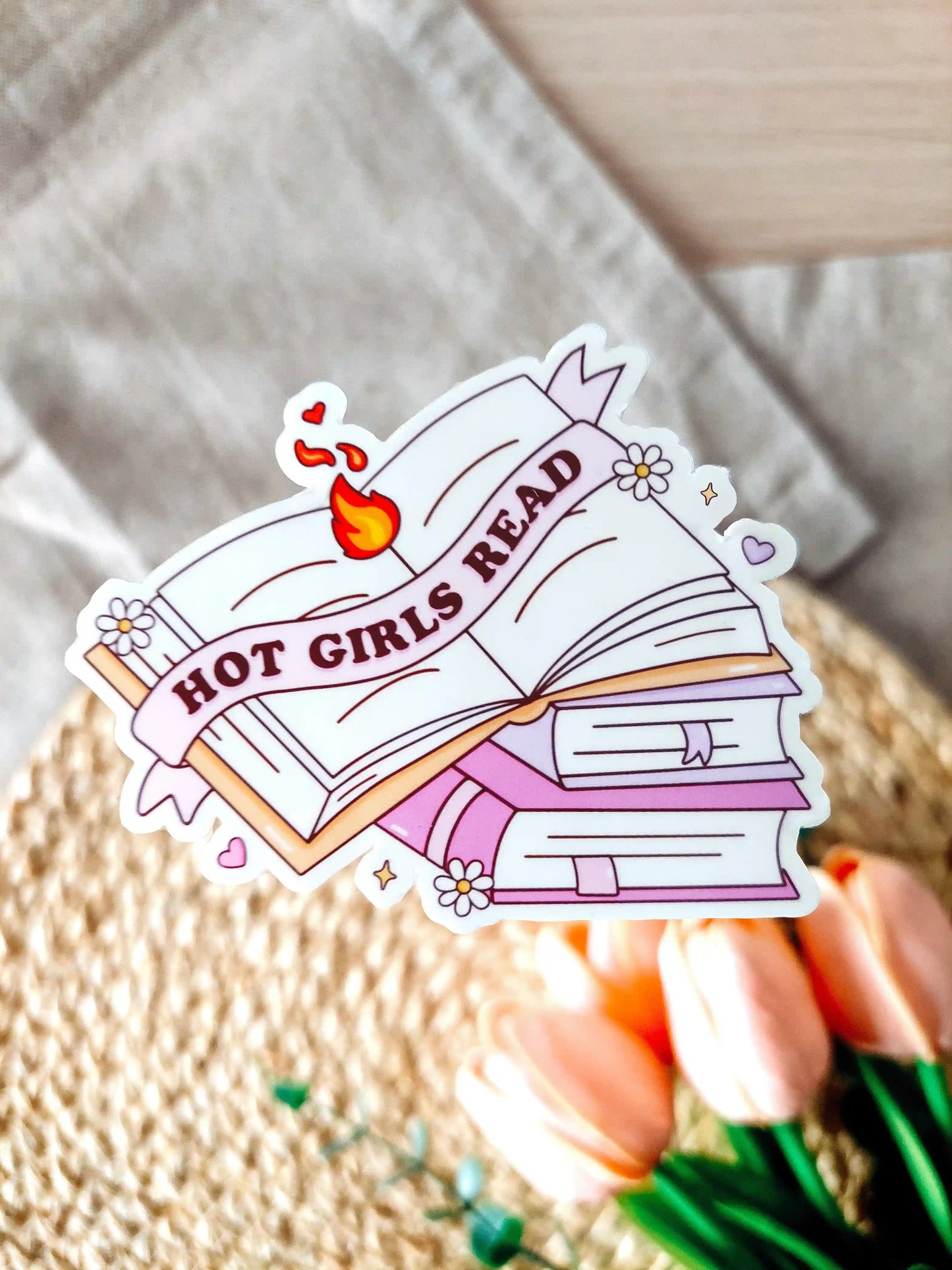 Vinyl sticker featuring an open book with a banner reading "Hot Girls Read" placed on top of a stack of books, photographed on a woven neutral background.