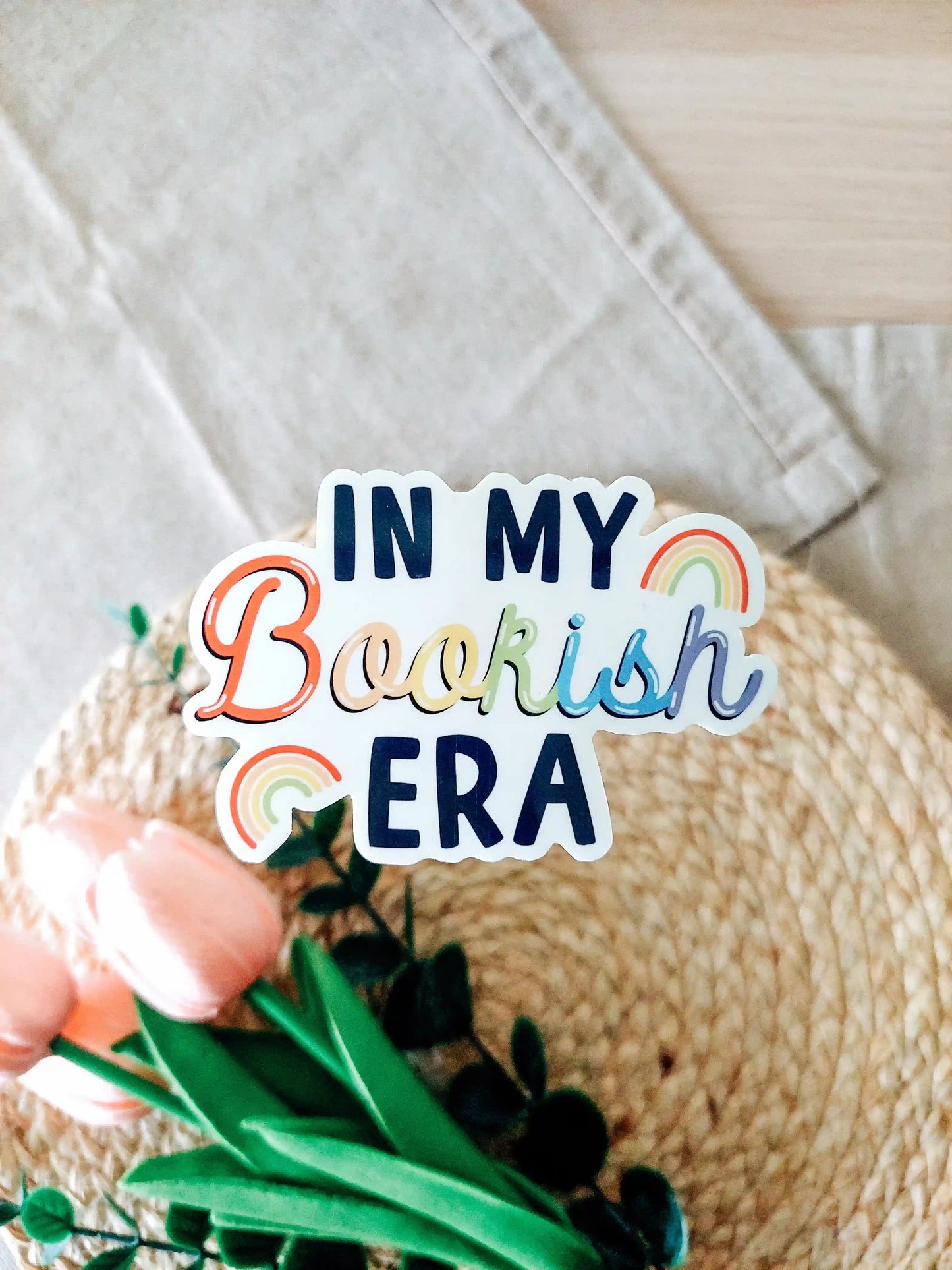 Vinyl sticker featuring the text "In My Bookish Era" in colorful rainbow lettering and small rainbow illustrations, photographed on neutral fabric background with woven mat.