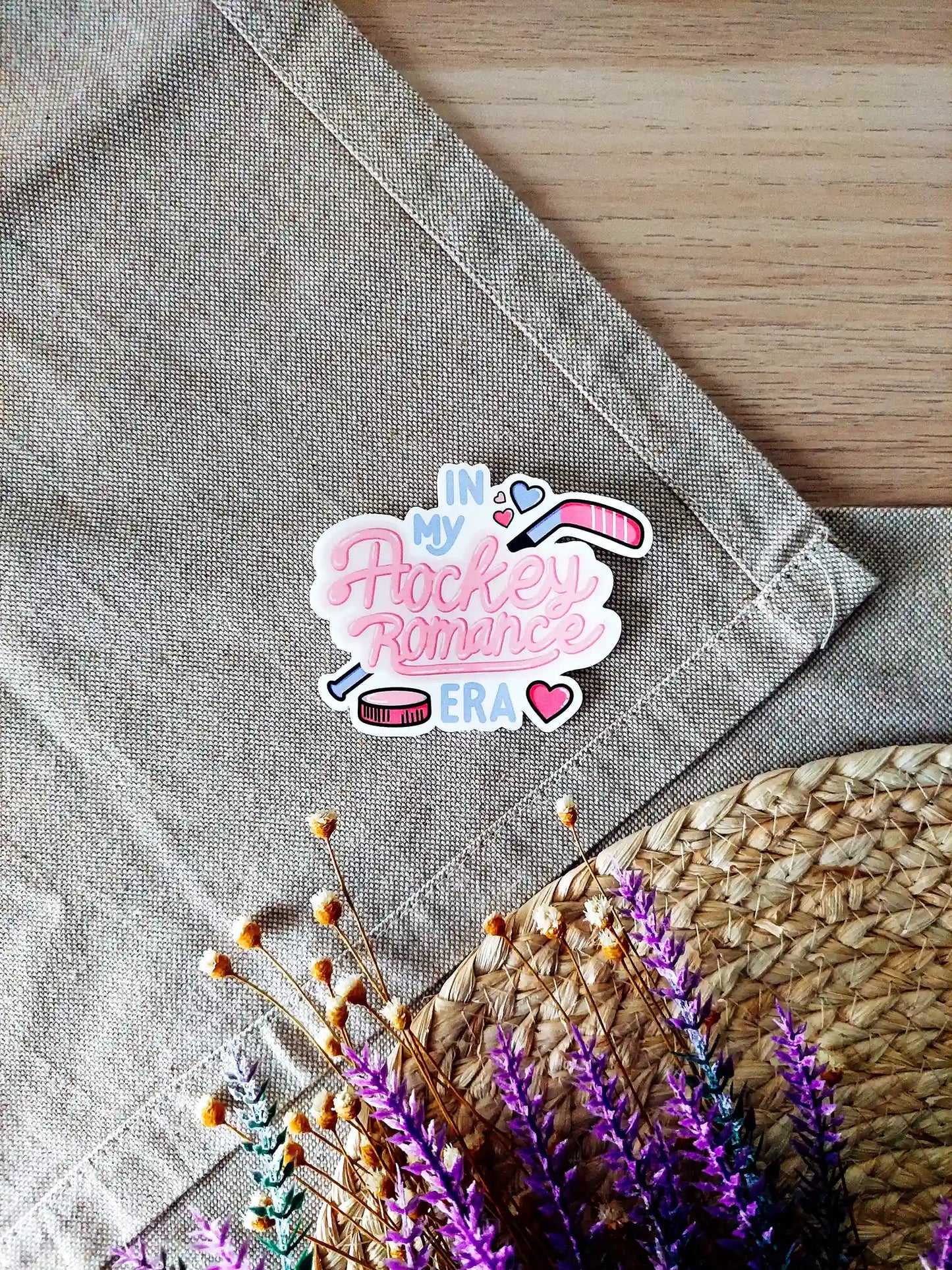 Vinyl sticker featuring the text "In My Hockey Romance Era" in pink lettering with hockey stick, puck and heart illustrations, photographed on neutral fabric background with woven mat.