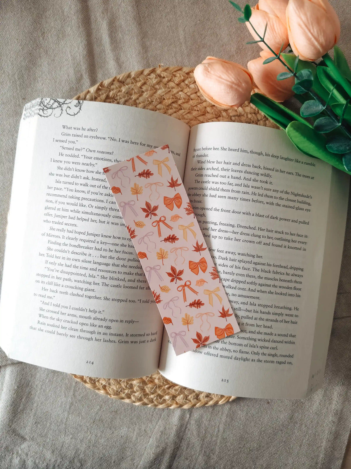 A laminated bookmark featuring scattered illustrated autumn leaves in shades of orange, red, and brown, and decorative bows in orange, yellow, and lavender on a pale blush background, resting on an open book on a woven rattan mat, with green foliage visible in the upper right corner.