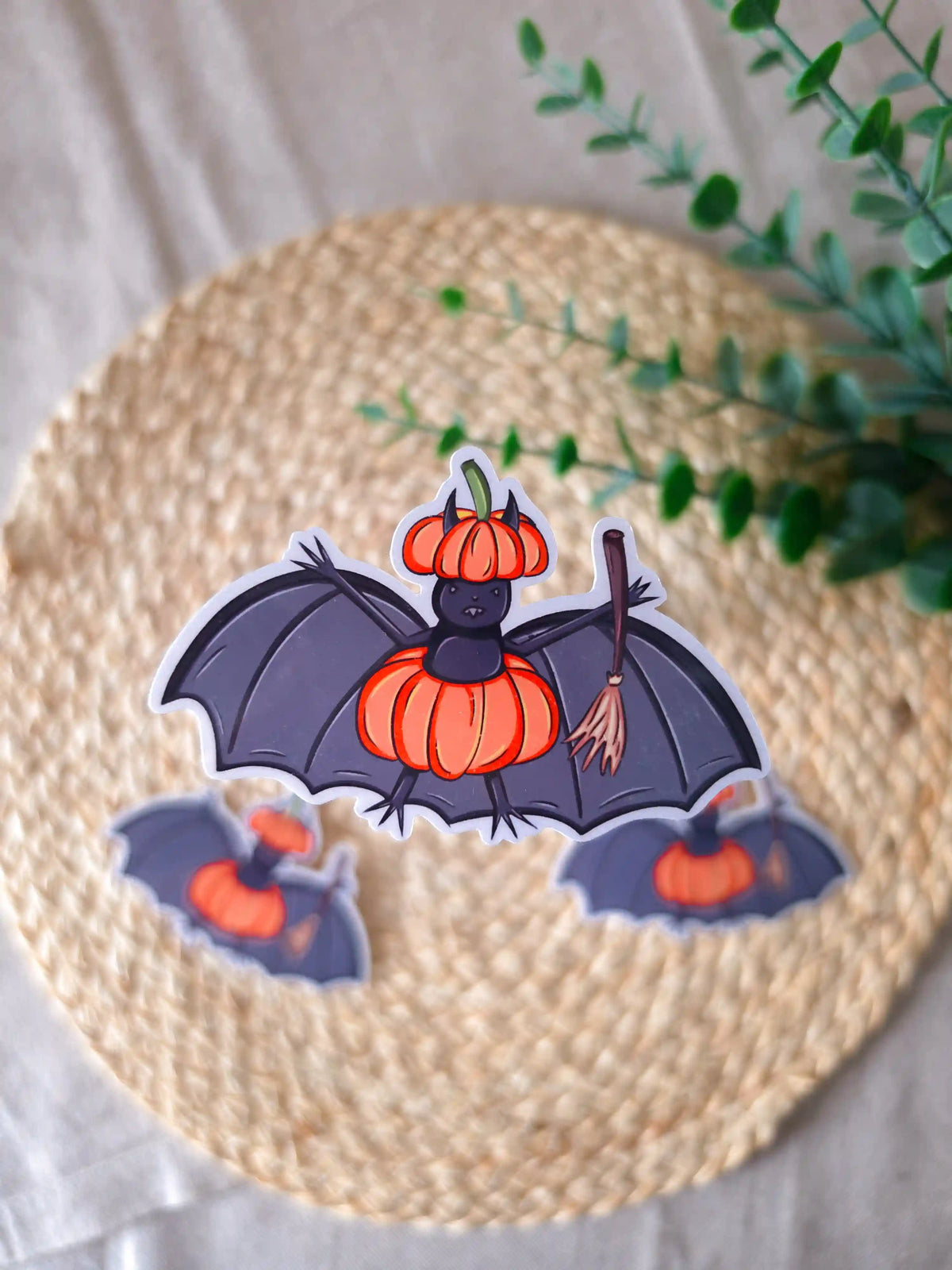 A large die-cut glossy vinyl sticker of a dark purple bat wearing an orange pumpkin as a costume, with a matching pumpkin-shaped hat and a wooden broomstick in one wing, displayed alongside two smaller versions of the same sticker on a woven rattan mat, with green eucalyptus sprigs in the upper right corner.