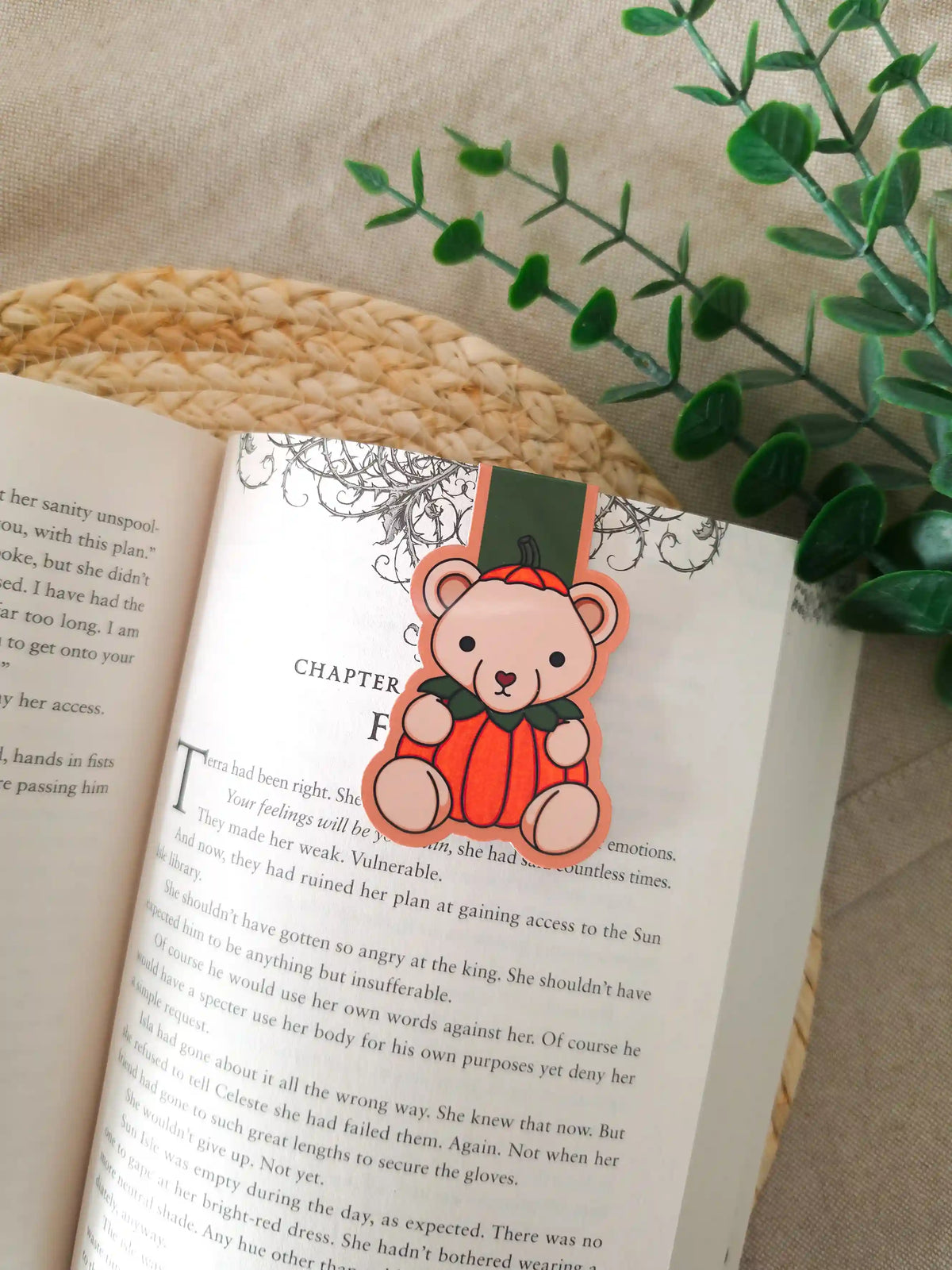 A die-cut magnetic bookmark featuring an illustrated beige teddy bear wearing a bright orange pumpkin costume with green leaves at the collar, clipped onto the top corner of an open book resting on a woven rattan mat, with green eucalyptus sprigs in the upper right corner.