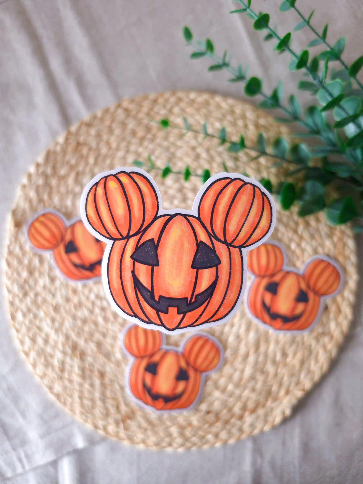 A large die-cut glossy vinyl sticker of a carved jack-o'-lantern pumpkin shaped like iconic round mouse ears, with a grinning carved face, displayed alongside two smaller versions of the same sticker on a woven rattan mat, with green eucalyptus sprigs in the upper right corner.