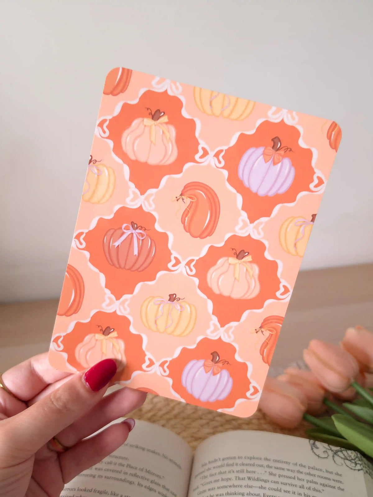 A laminated e-reader insert featuring a warm orange and terracotta tile pattern with illustrated pumpkins in peach, lavender, and blush tones, some decorated with small bows, resting on an open book on a woven rattan mat, with green eucalyptus sprigs in the upper right corner.
