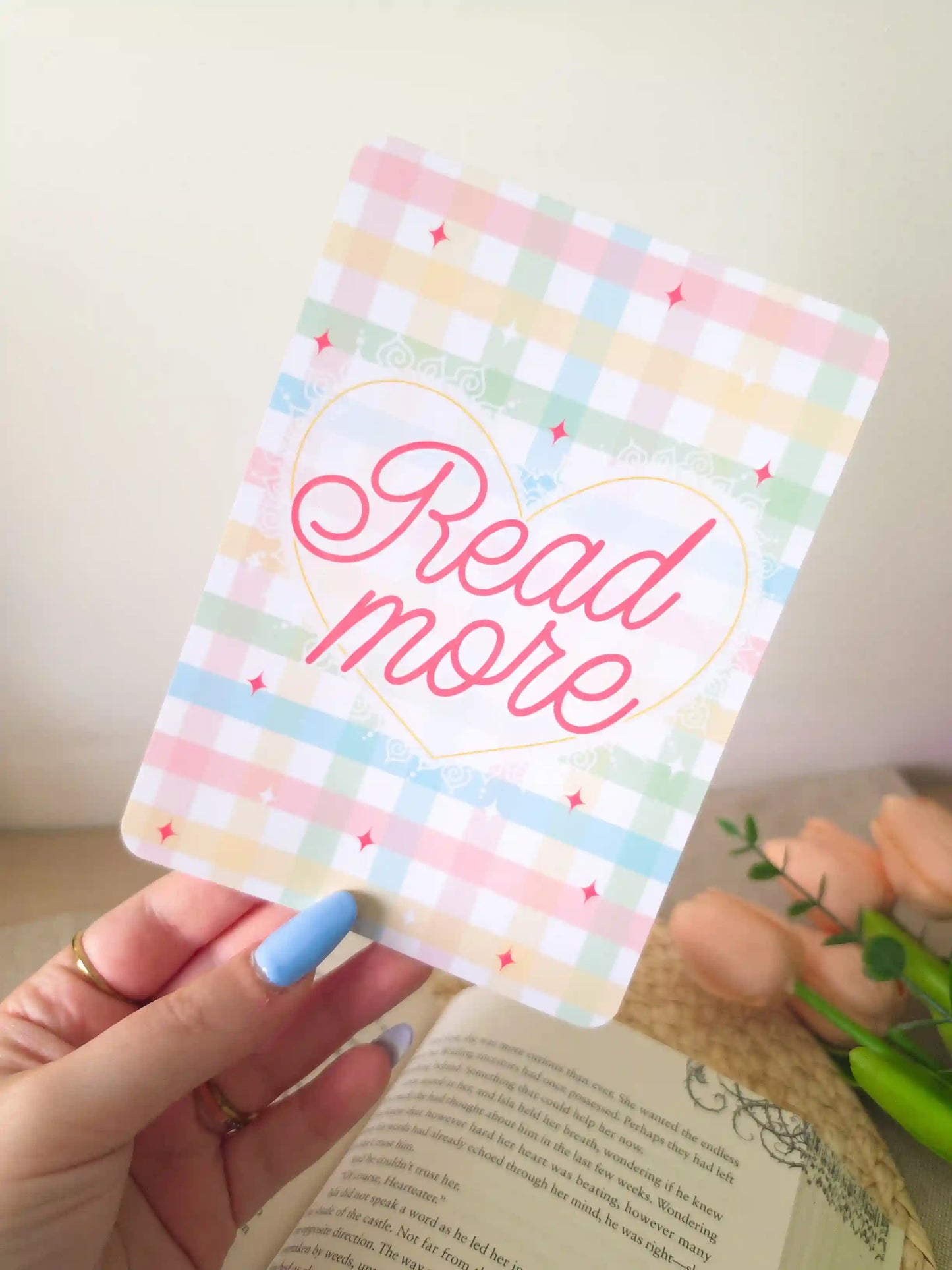 A hand holding an e-reader insert with a pastel gingham background in soft pink, blue, green, and yellow, featuring a gold heart outline with the text "Read More" in pink cursive script and pink sparkle details.