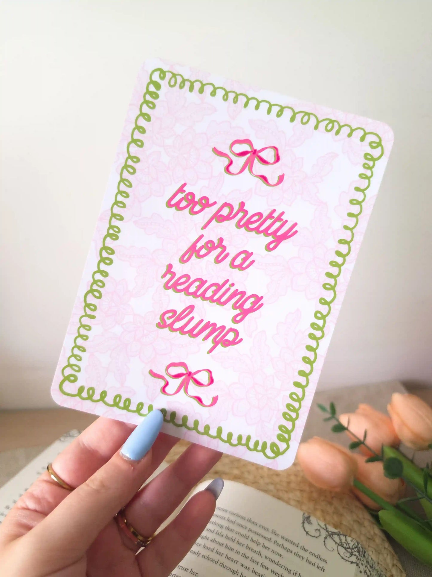 A hand holding an e-reader insert with a pink lacey background, green wavy decorative border, two pink bow details, and the text "too pretty for a reading slump" in bold pink cursive script.