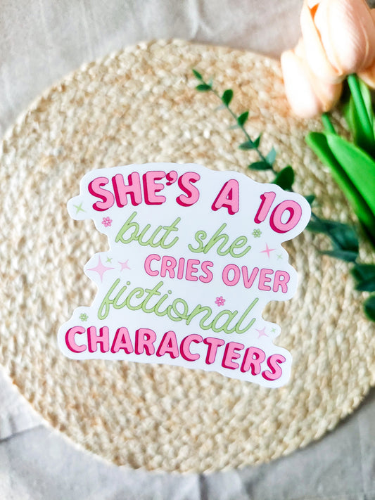 "She's A 10 But She Cries Over Fictional Characters" sticker on a rattan placemat, featuring pink and green text with small flowers and stars detail.