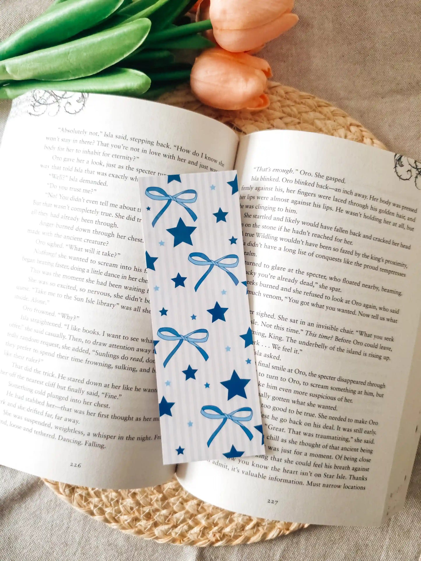 An open book spread on a woven mat with green foliage around it. A decorative bookmark with blue ribbon bows and stars is placed across the pages.