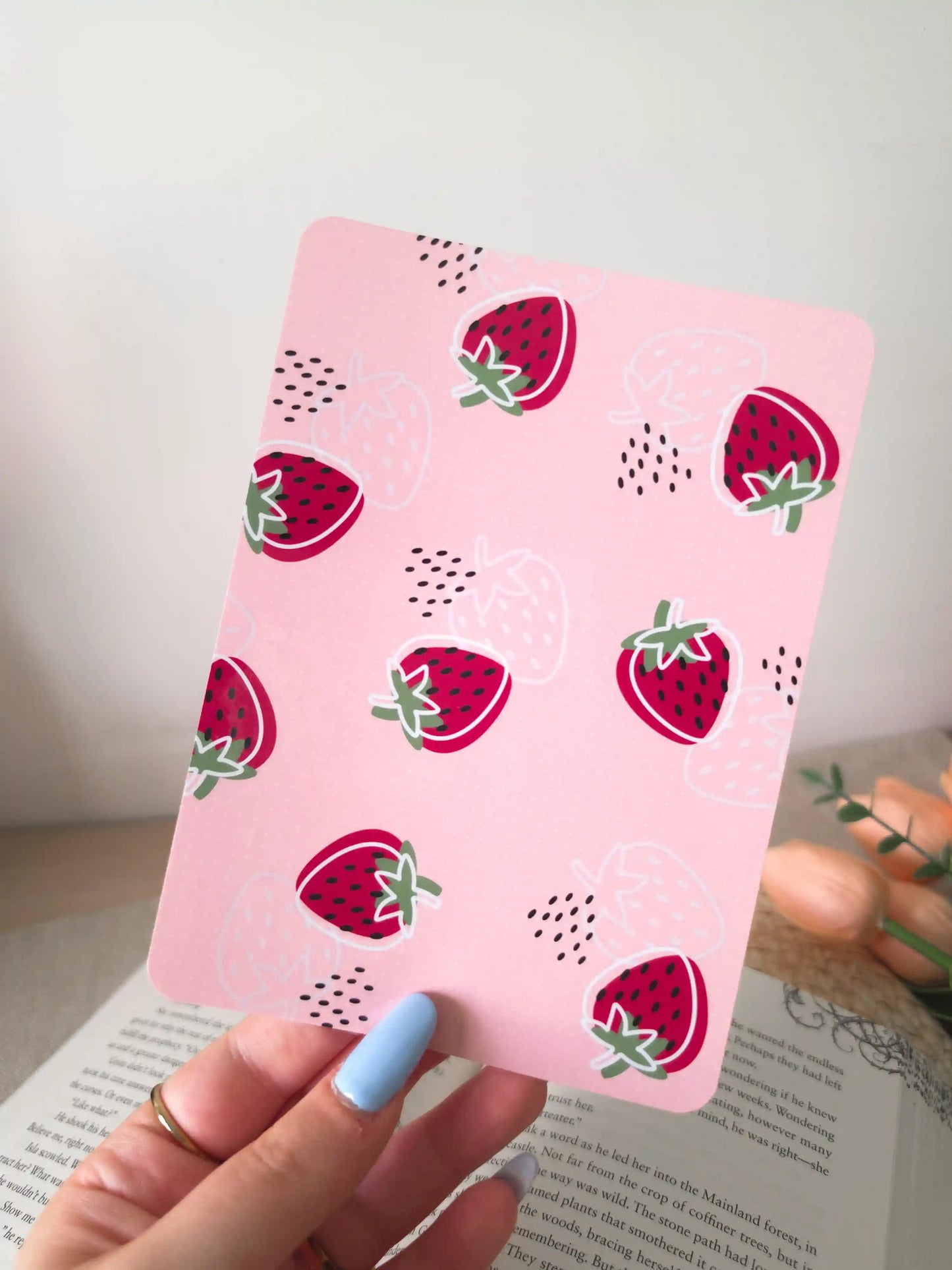 A hand holding an e-reader insert with a solid pink background featuring red strawberry illustrations with white outlines and green leaves, with small black dot details scattered across it.