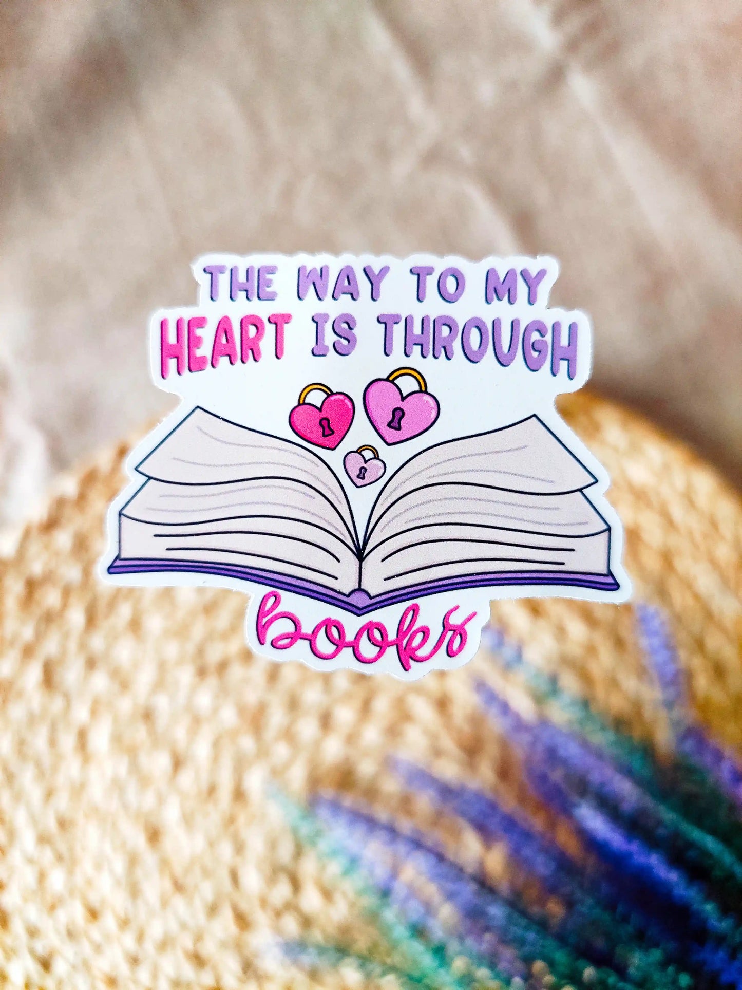 Vinyl sticker featuring an open book with pink heart-shaped locks and the text "The way to my heart is through books", photographed on a neutral fabric background with woven mat.