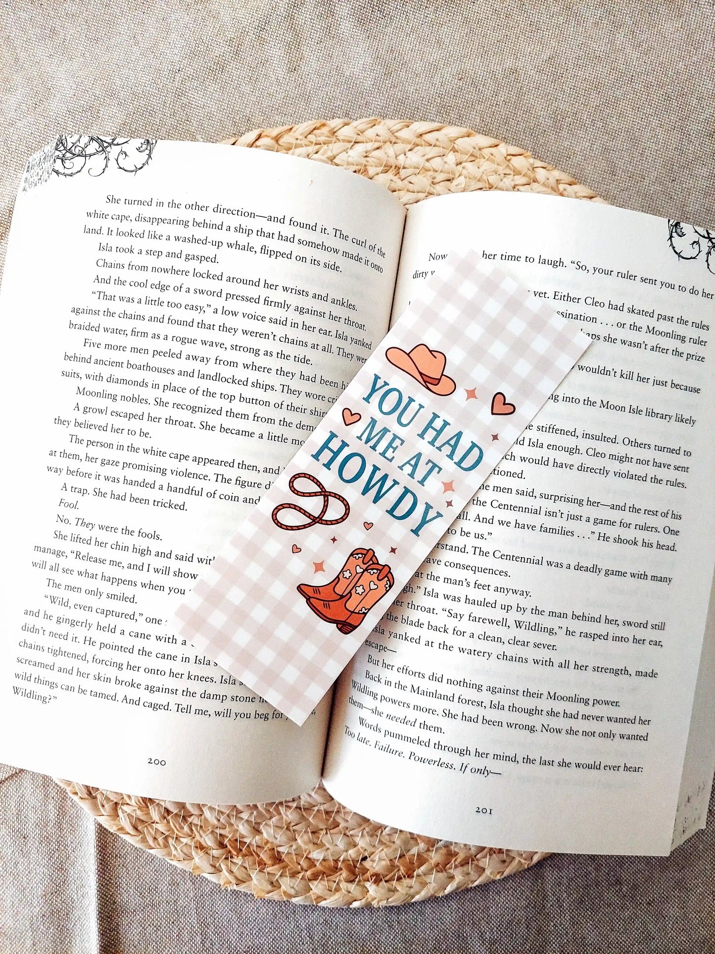 Bookmark with the text "You Had Me at Howdy", featuring a western-themed design with a cowboy hat, boots and rope on a soft gingham background, resting on an open book.