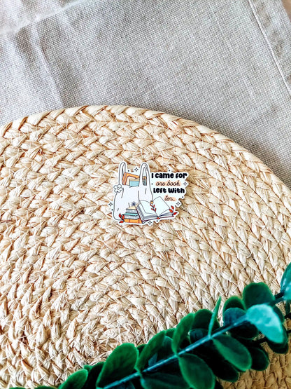Vinyl bookish sticker featuring a tote bag filled with books and the phrase "I came for one book, left with ten", photographed on a neutral textured background.