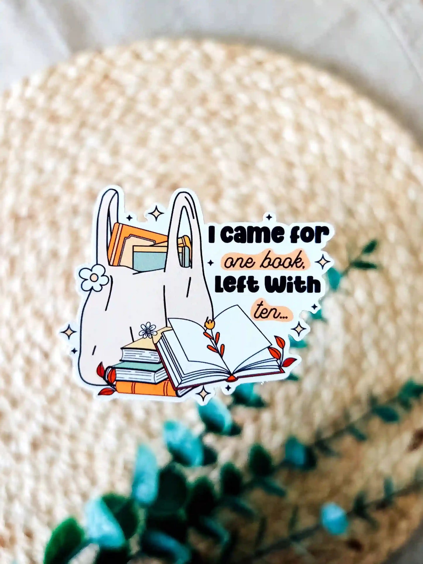 Vinyl bookish sticker featuring a tote bag filled with books and the phrase "I came for one book, left with ten", photographed on a neutral textured background.