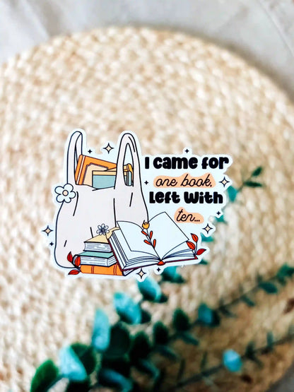 Vinyl bookish sticker featuring a tote bag filled with books and the phrase "I came for one book, left with ten", photographed on a neutral textured background.