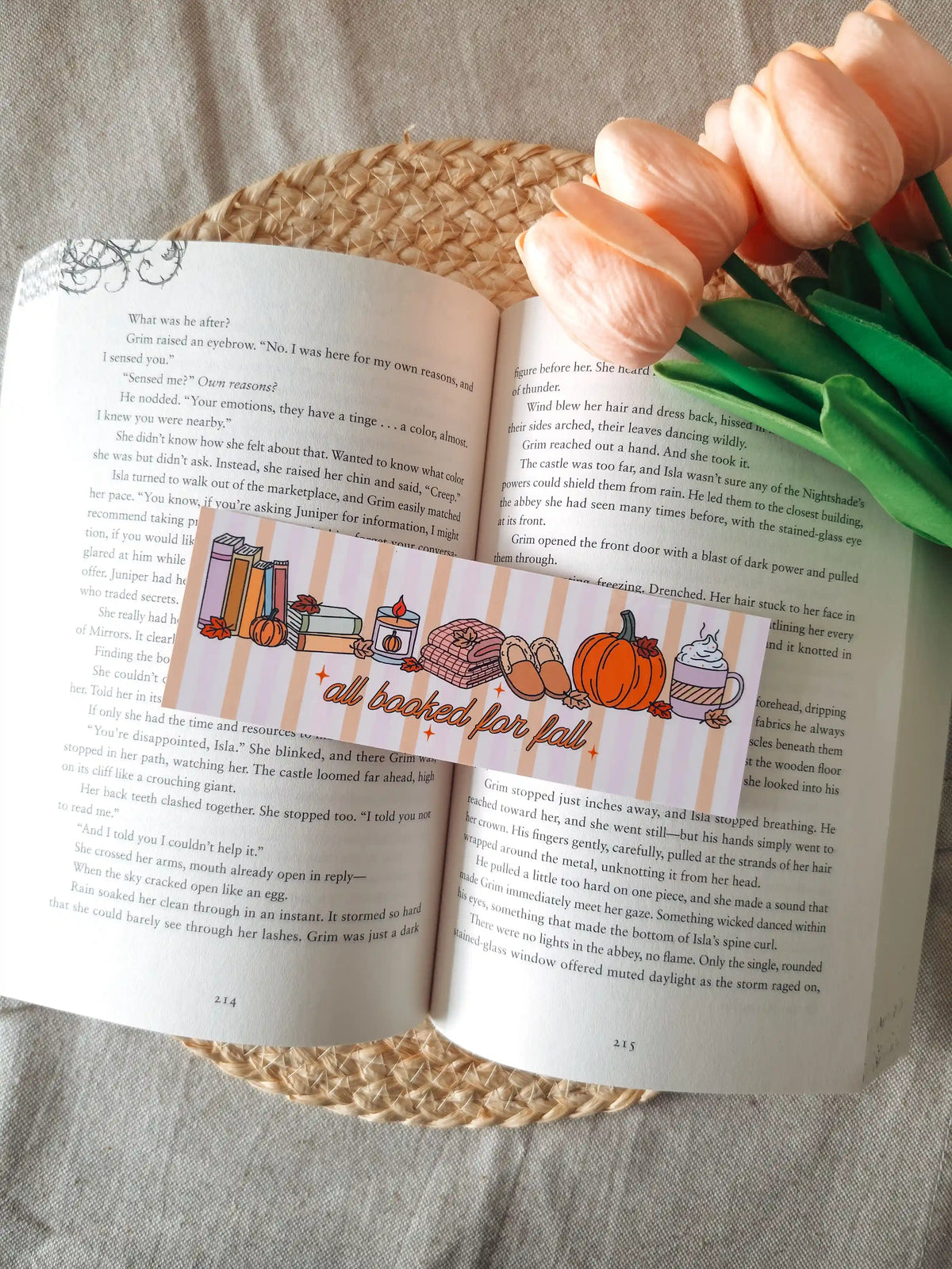 A horizontal laminated bookmark with a pastel striped background featuring the text "all booked for fall" in orange cursive lettering, decorated with illustrated autumn items including a stack of books, a lit candle, a knit blanket, slippers, a pumpkin, and a whipped cream latte, resting on an open book on a woven rattan mat, with peach tulips in the upper right corner.