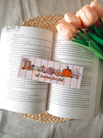 A horizontal laminated bookmark with a pastel striped background featuring the text "all booked for fall" in orange cursive lettering, decorated with illustrated autumn items including a stack of books, a lit candle, a knit blanket, slippers, a pumpkin, and a whipped cream latte, resting on an open book on a woven rattan mat, with peach tulips in the upper right corner.