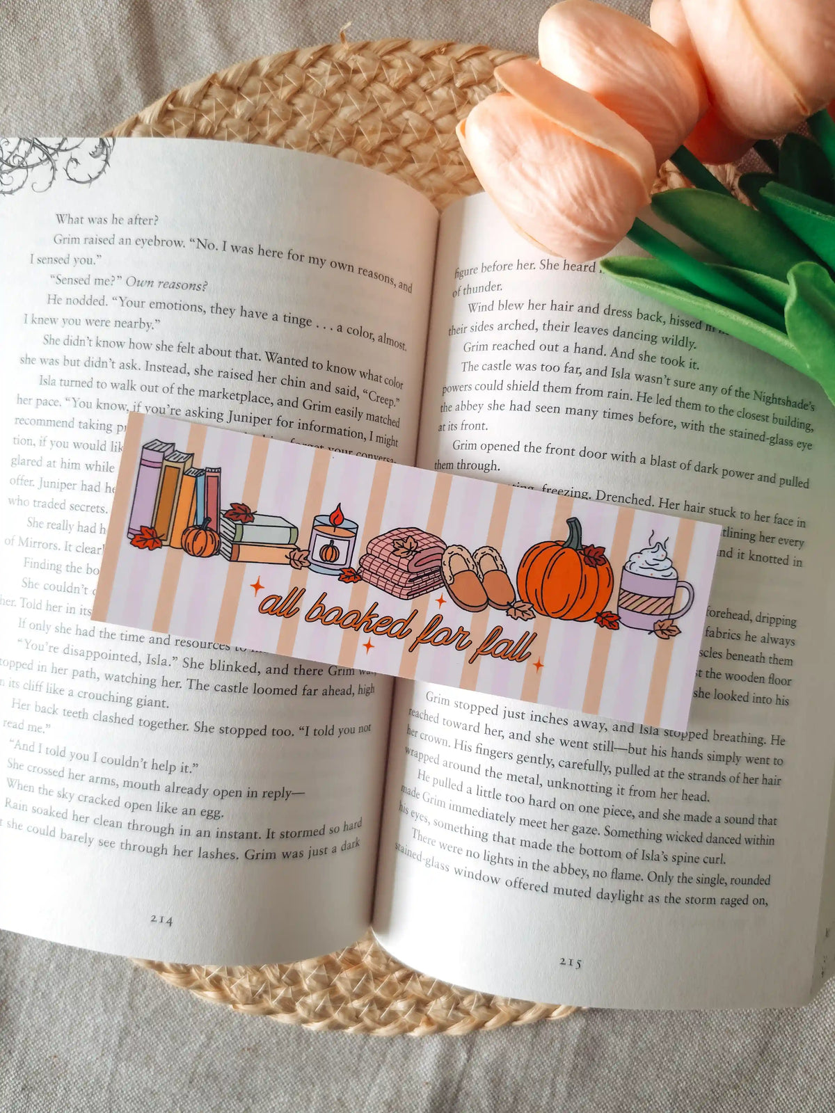 A horizontal laminated bookmark with a pastel striped background featuring the text "all booked for fall" in orange cursive lettering, decorated with illustrated autumn items including a stack of books, a lit candle, a knit blanket, slippers, a pumpkin, and a whipped cream latte, resting on an open book on a woven rattan mat, with peach tulips in the upper right corner.