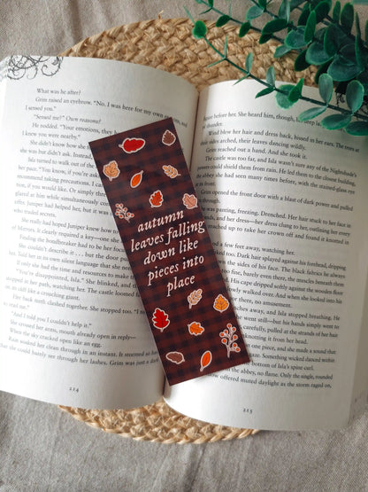 A laminated bookmark on a dark brown gingham background featuring the text "autumn leaves falling down like pieces into place" in white italic lettering, surrounded by illustrated autumn leaves in red, orange, and brown, and a red berry branch, resting on an open book on a woven rattan mat, with green eucalyptus sprigs in the upper right corner.