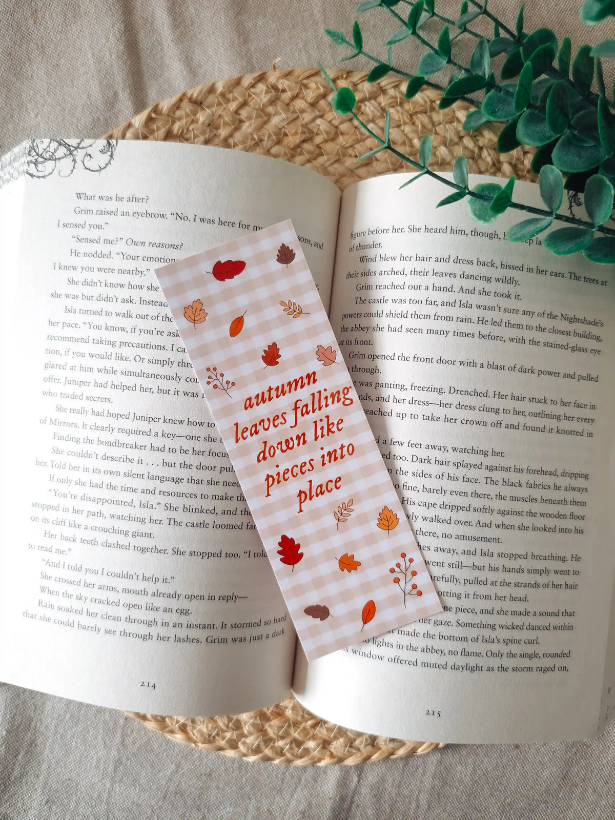 A laminated bookmark on a pale pink and white gingham background featuring the text "autumn leaves falling down like pieces into place" in red italic lettering, surrounded by illustrated autumn leaves in red, orange, and brown, and a small berry branch, resting on an open book on a woven rattan mat, with green eucalyptus sprigs in the upper right corner.