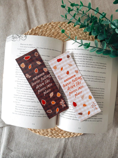 Two laminated bookmarks resting on an open book on a woven rattan mat: one on a brown gingham background with white lettering and one on a pale pink gingham background with red lettering, both featuring the text "autumn leaves falling down like pieces into place" and illustrated autumn leaves and berry branches, with green eucalyptus sprigs in the upper right corner.