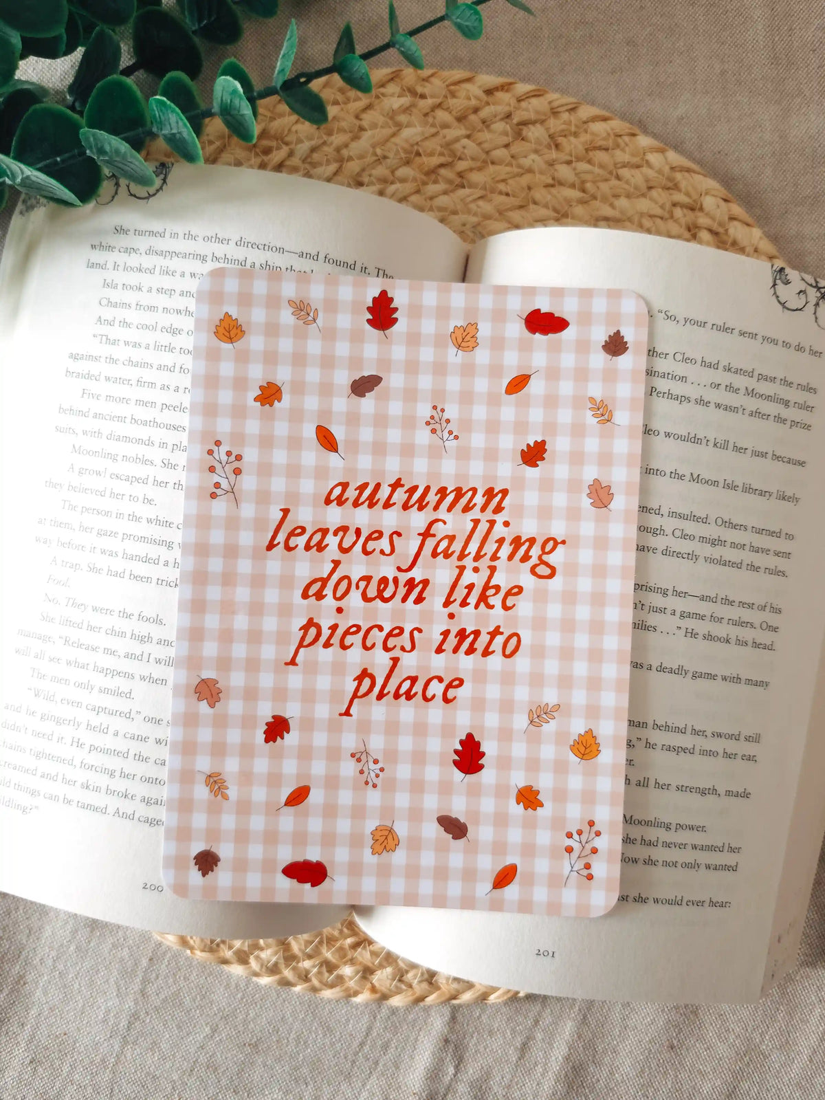 A laminated e-reader insert on a pale pink gingham background featuring the text "autumn leaves falling down like pieces into place" in orange-red cursive lettering at the centre, surrounded by illustrated autumn leaves in shades of red, orange, and brown, and small berry branches, resting on an open book on a woven rattan mat, with green eucalyptus branches visible in the upper left corner.