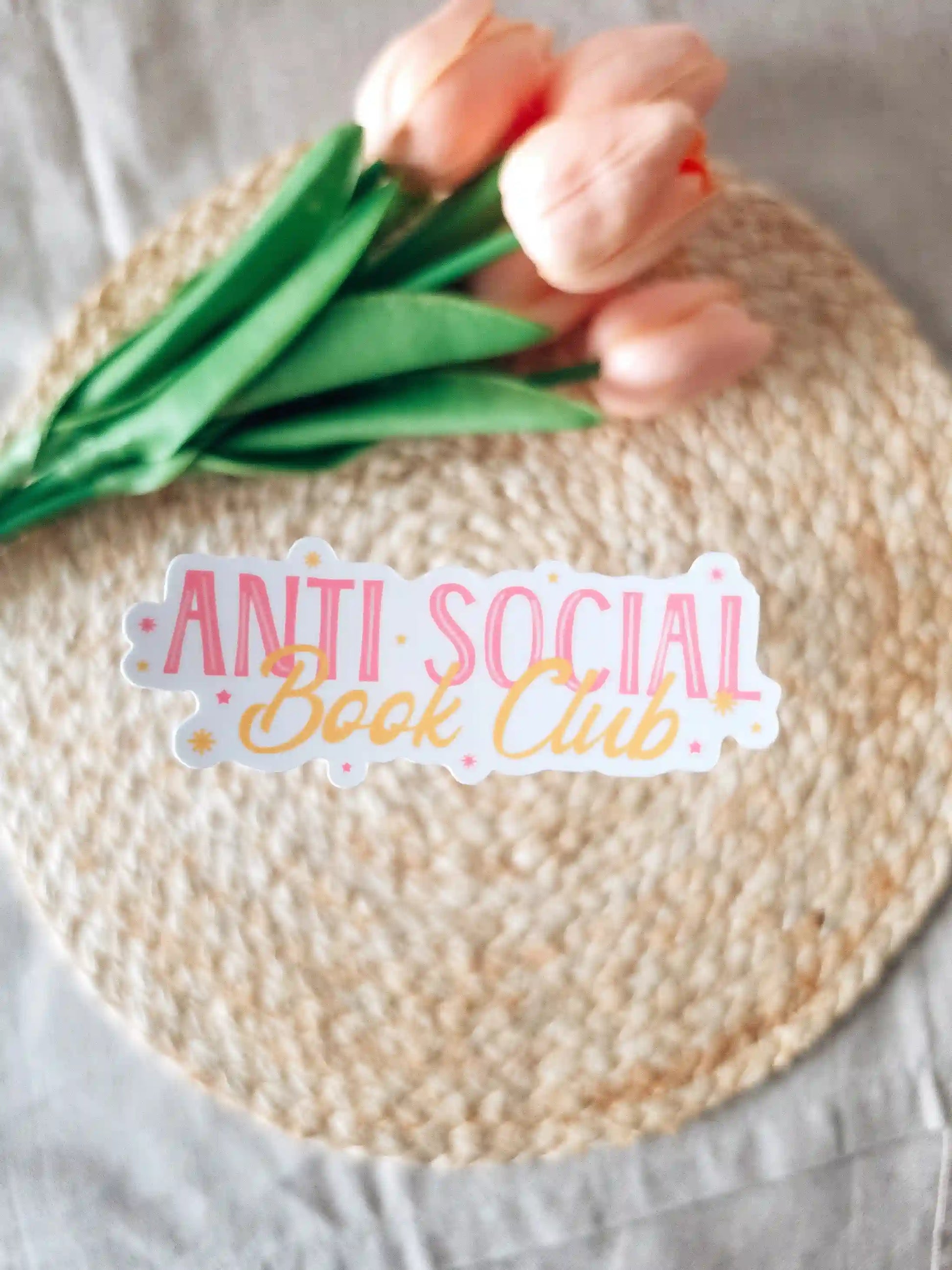 A die-cut sticker reading "Anti Social Book Club" in pink block lettering and gold cursive script, with small star details on a white background, placed on a woven rattan mat with peach roses and green leaves resting in the upper corner.