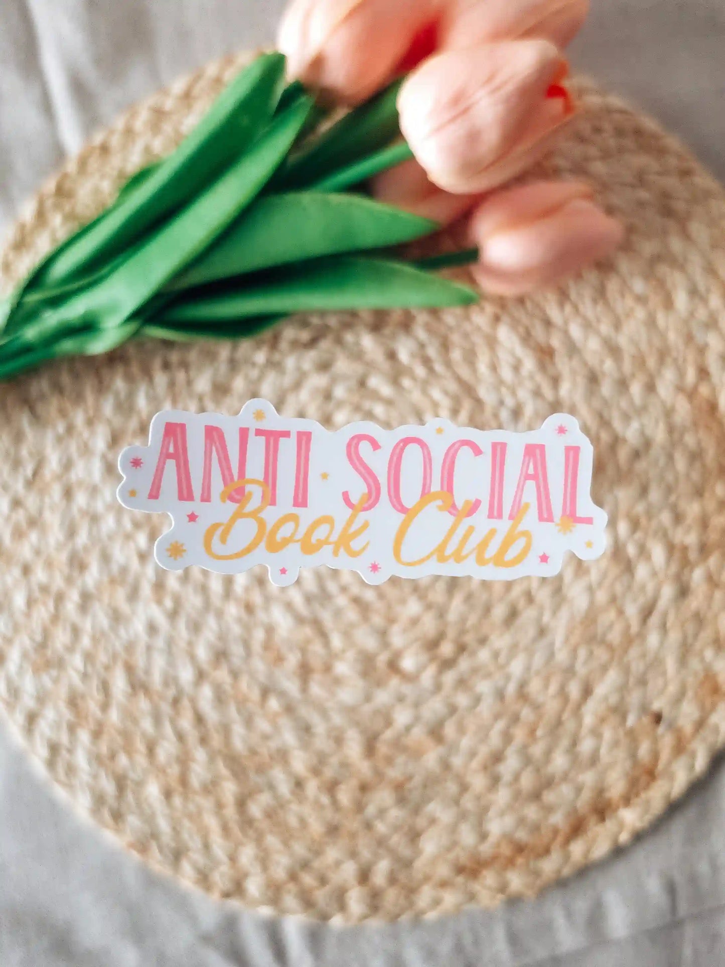 A die-cut sticker reading "Anti Social Book Club" in pink block lettering and gold cursive script, with small star details on a white background, placed on a woven rattan mat with peach roses and green leaves resting in the upper corner.