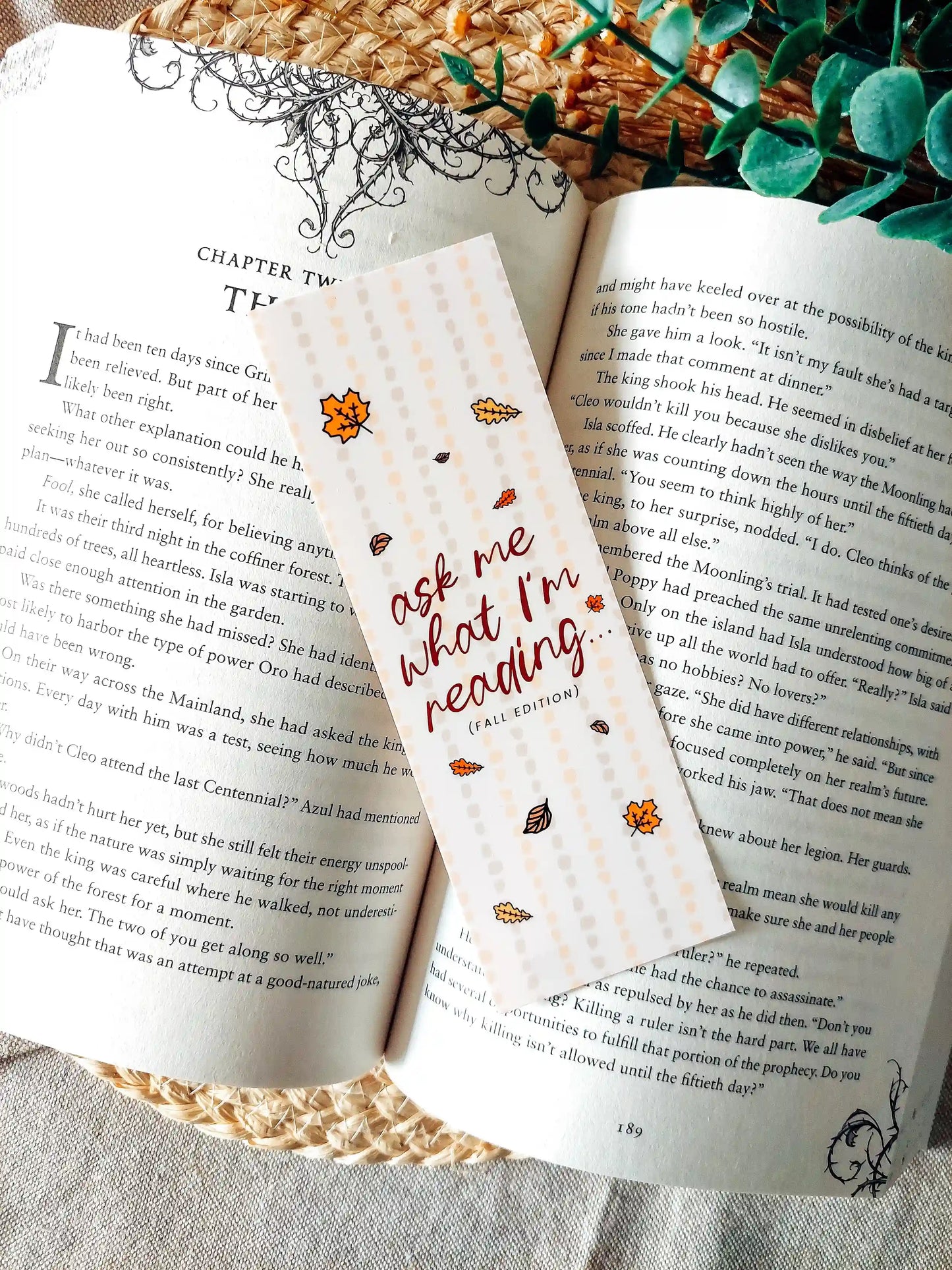 Illustrated bookmark in warm autumn colours with the text "Ask Me What I’m Reading" decorated with autumn leaves and placed on an open book.