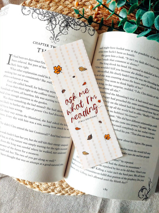 Illustrated bookmark in warm autumn colours with the text "Ask Me What I’m Reading" decorated with autumn leaves and placed on an open book.