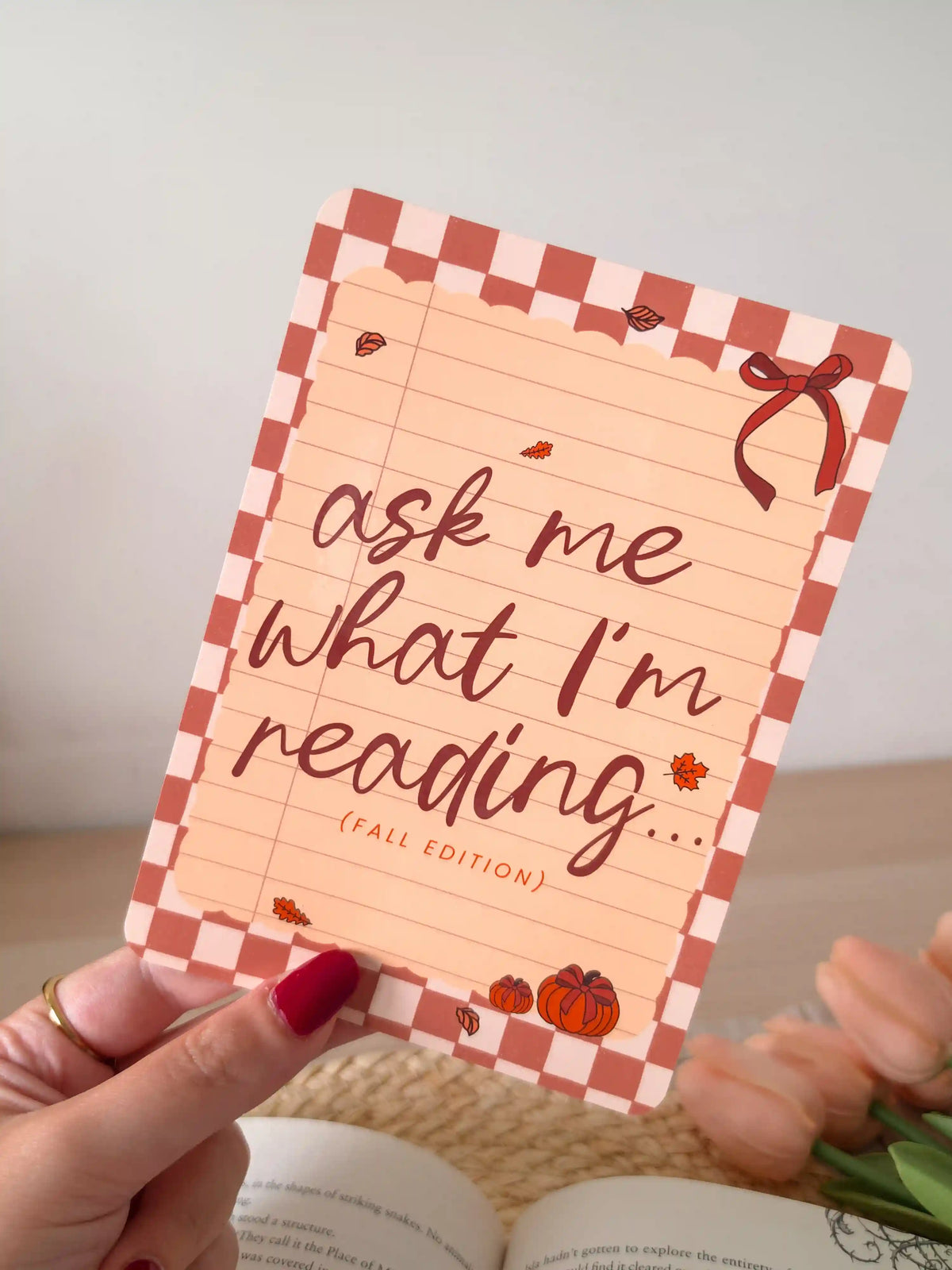 A laminated e-reader insert with a terracotta and blush checkered border, a lined notebook-style peach center, the text "ask me what I'm reading... (fall edition)" in dark brown cursive lettering, a red ribbon bow in the upper right corner, and small illustrated autumn leaves and pumpkins, resting on an open book on a woven rattan mat, with green leafy branches in the upper left corner.
