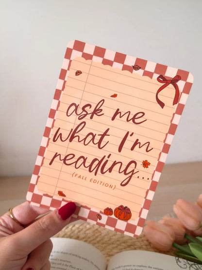 A laminated e-reader insert with a terracotta and blush checkered border, a lined notebook-style peach center, the text "ask me what I'm reading... (fall edition)" in dark brown cursive lettering, a red ribbon bow in the upper right corner, and small illustrated autumn leaves and pumpkins, resting on an open book on a woven rattan mat, with green leafy branches in the upper left corner.