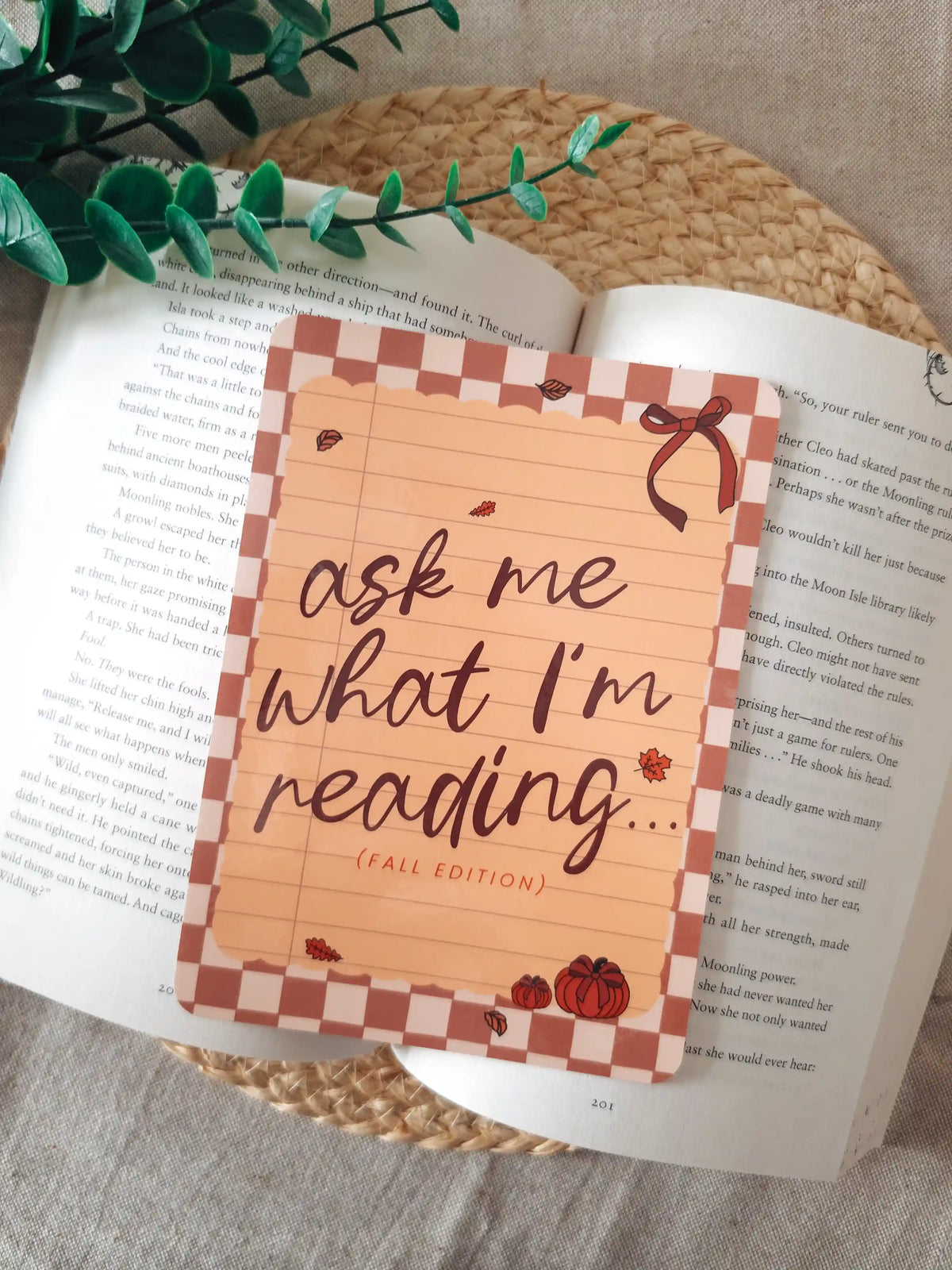 A laminated e-reader insert with a terracotta and blush checkered border, a lined notebook-style peach center, the text "ask me what I'm reading... (fall edition)" in dark brown cursive lettering, a red ribbon bow in the upper right corner, and small illustrated autumn leaves and pumpkins, resting on an open book on a woven rattan mat, with green leafy branches in the upper left corner.