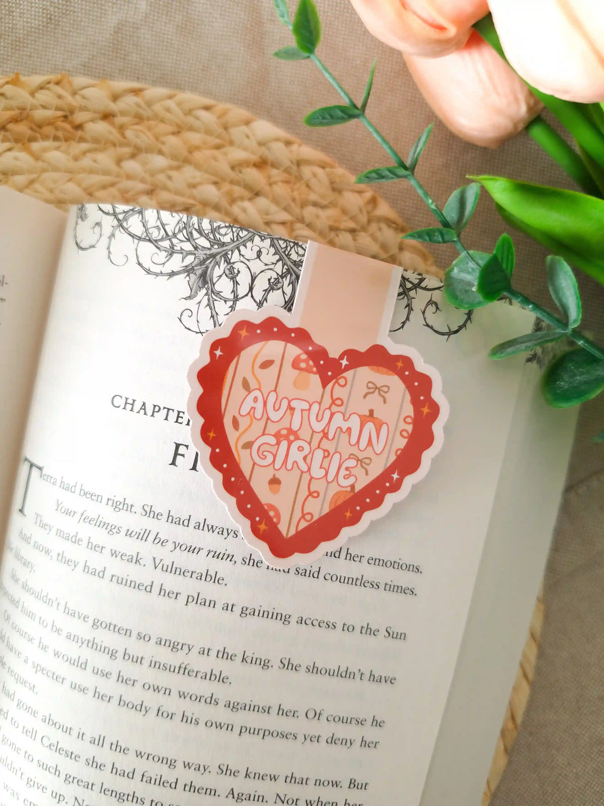 A die-cut heart-shaped magnetic bookmark with a scalloped dark red border, a peach striped background, the text "Autumn Girlie" in pink lettering at the centre, and small illustrated autumn details including a mushroom, a bow, an acorn, and sparkles, clipped onto the top corner of an open book resting on a woven rattan mat, with peach tulips and green eucalyptus sprigs in the upper right corner.
