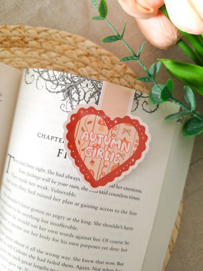 A die-cut heart-shaped magnetic bookmark with a scalloped dark red border, a peach striped background, the text "Autumn Girlie" in pink lettering at the centre, and small illustrated autumn details including a mushroom, a bow, an acorn, and sparkles, clipped onto the top corner of an open book resting on a woven rattan mat, with peach tulips and green eucalyptus sprigs in the upper right corner.