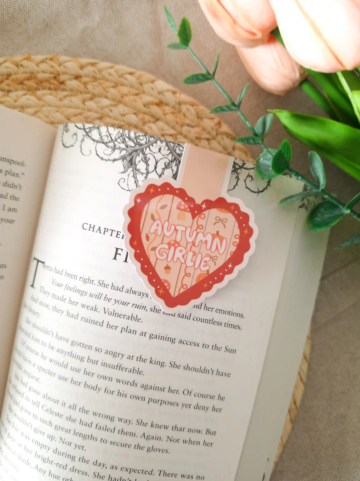 A die-cut heart-shaped magnetic bookmark with a scalloped dark red border, a peach striped background, the text "Autumn Girlie" in pink lettering at the centre, and small illustrated autumn details including a mushroom, a bow, an acorn, and sparkles, clipped onto the top corner of an open book resting on a woven rattan mat, with peach tulips and green eucalyptus sprigs in the upper right corner.