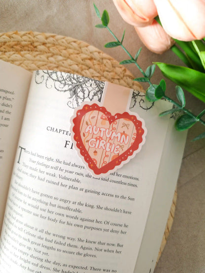 A die-cut heart-shaped magnetic bookmark with a scalloped dark red border, a peach striped background, the text "Autumn Girlie" in pink lettering at the centre, and small illustrated autumn details including a mushroom, a bow, an acorn, and sparkles, clipped onto the top corner of an open book resting on a woven rattan mat, with peach tulips and green eucalyptus sprigs in the upper right corner.