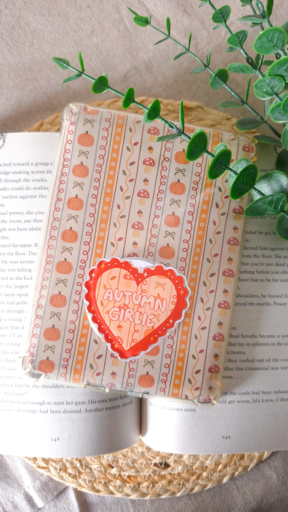 Orange heart magnetic grip with the text “Autumn Girlie” attached to an e-reader with an autumn insert of pumpkins, mushrooms, bows and acorns, lying on an open book on a woven rattan mat, with a green eucalyptus sprig across the corner.