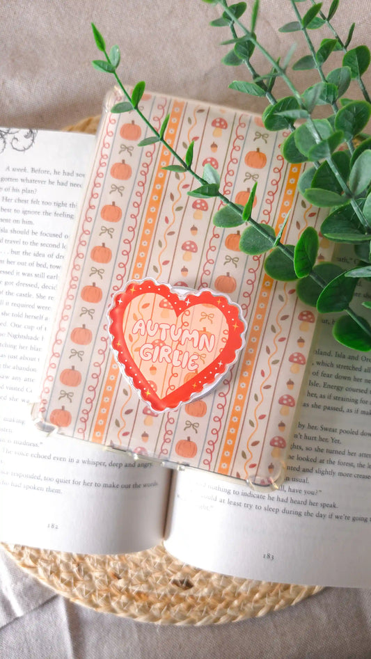 Orange heart magnetic grip with the text “Autumn Girlie” attached to an e-reader with an autumn insert of pumpkins, mushrooms, bows and acorns, lying on an open book on a woven rattan mat, with a green eucalyptus sprig across the corner.