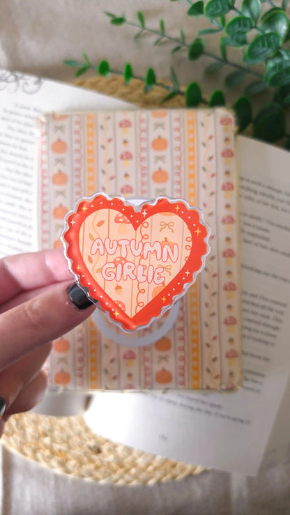 Hand holding an orange heart-shaped acrylic grip with the text “Autumn Girlie” over a pattern of pumpkins, mushrooms and bows, in front of an autumn-patterned magnetic bookmark, an open book, a green plant and a woven rattan mat.
